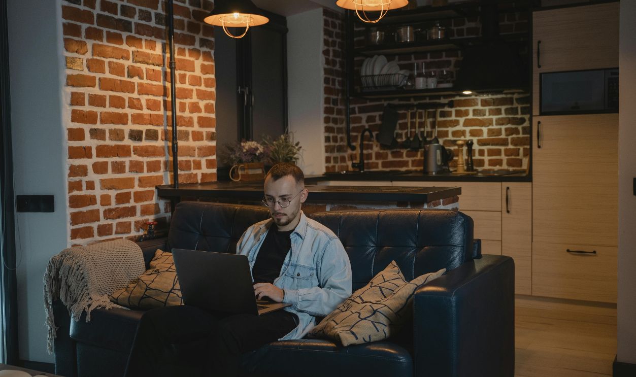 A bearded man sits comfortably on a couch, working on his laptop in a stylish living room