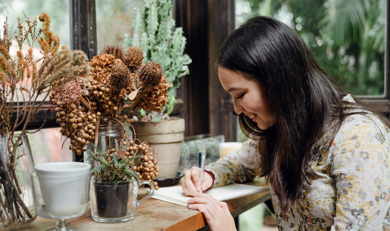 A cheerful young woman writes in a notebook surrounded by indoor plants, creating a cozy and creative workspace