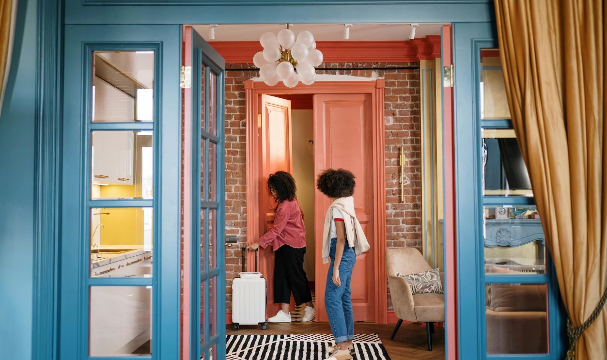 Stylish room with two women walking through doorway, carrying suitcase. Cozy and colorful decor