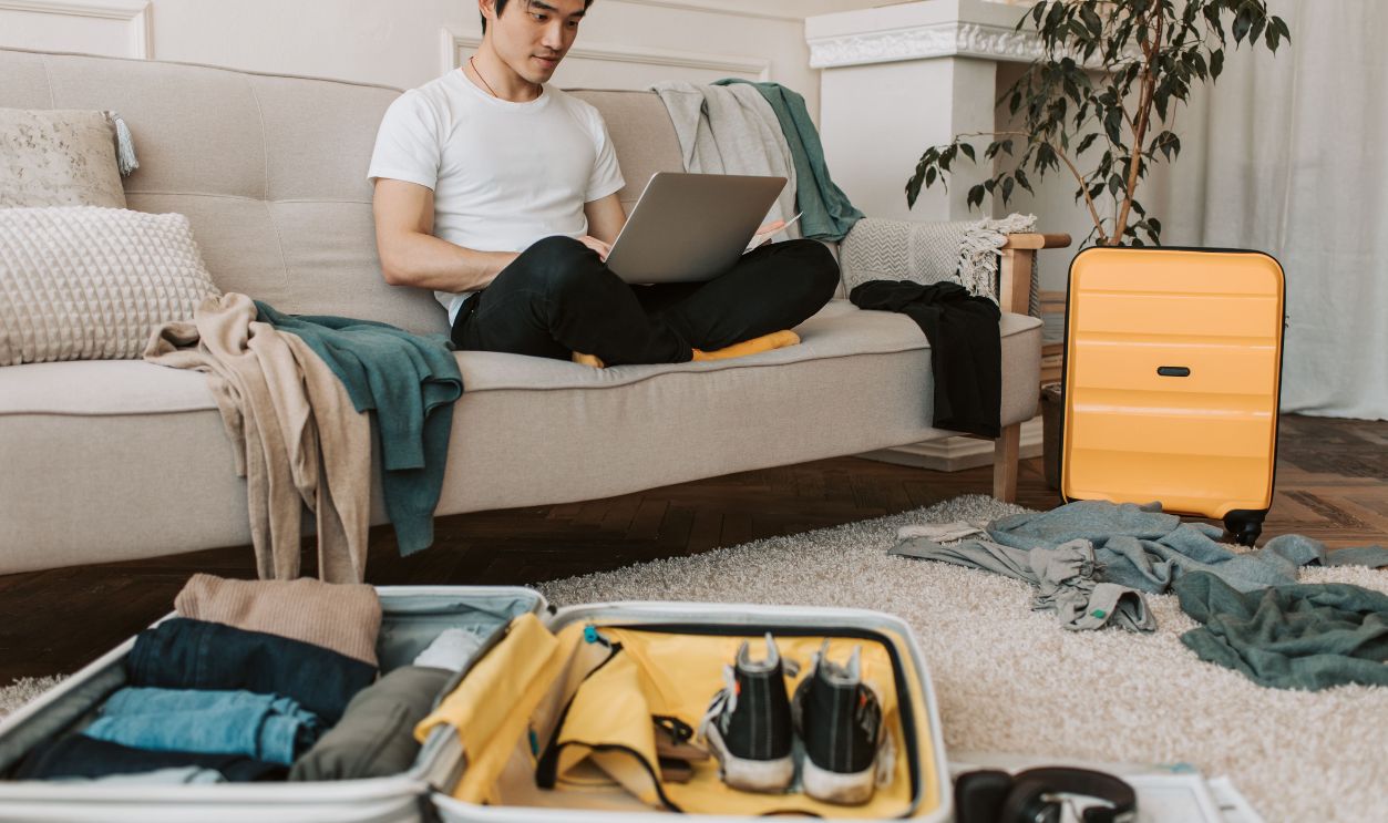 A young man in yellow accessories works on a laptop among unpacked luggage at home