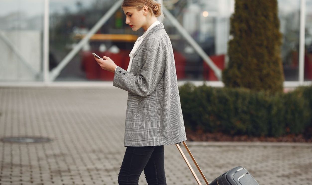 Stylish woman walking with suitcase while using smartphone outside a modern building.