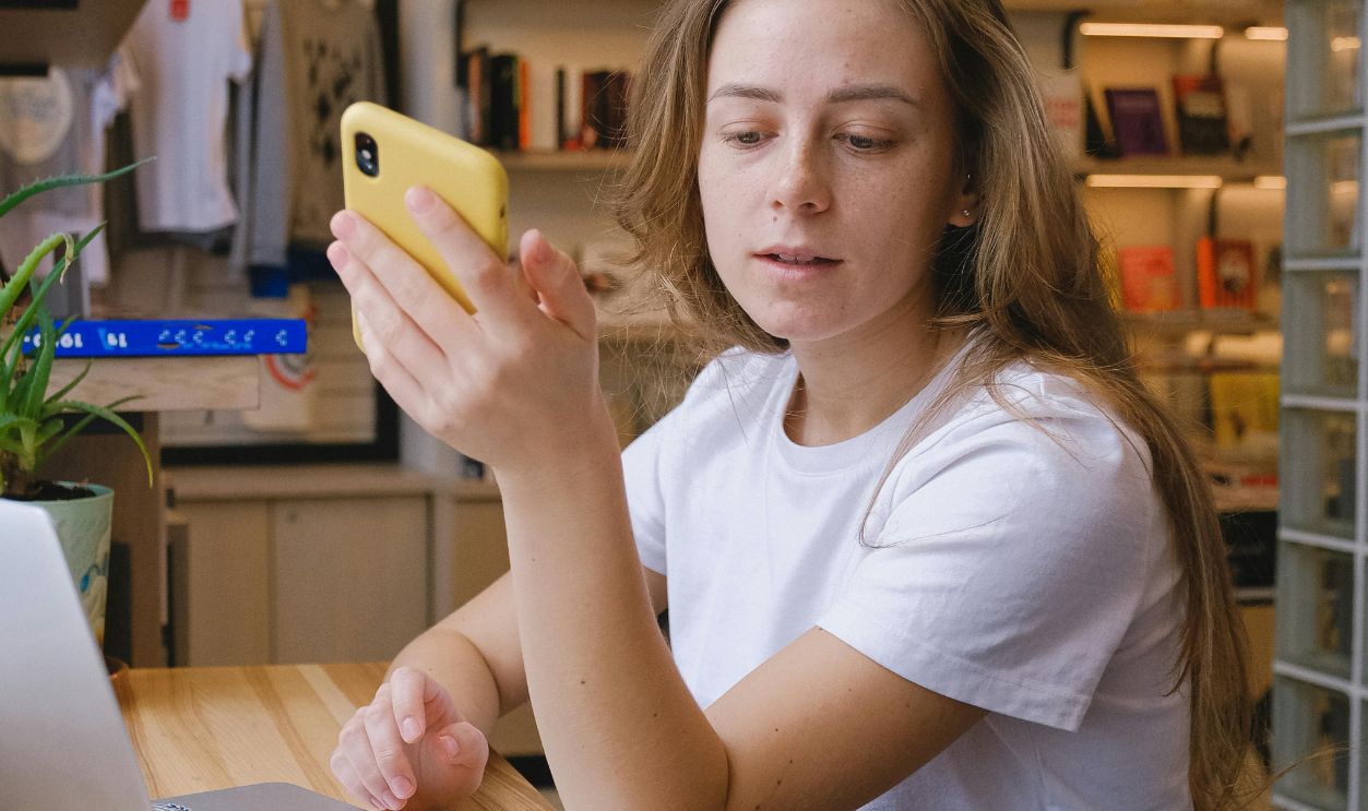 Young woman sitting in a bookstore, using a smartphone by her laptop, surrounded by books