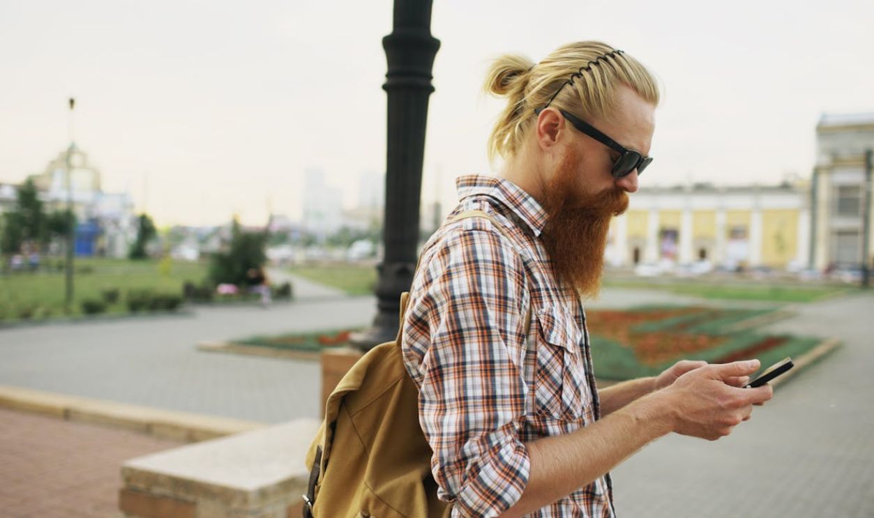 Bearded man in sunglasses and plaid shirt using smartphone outdoors