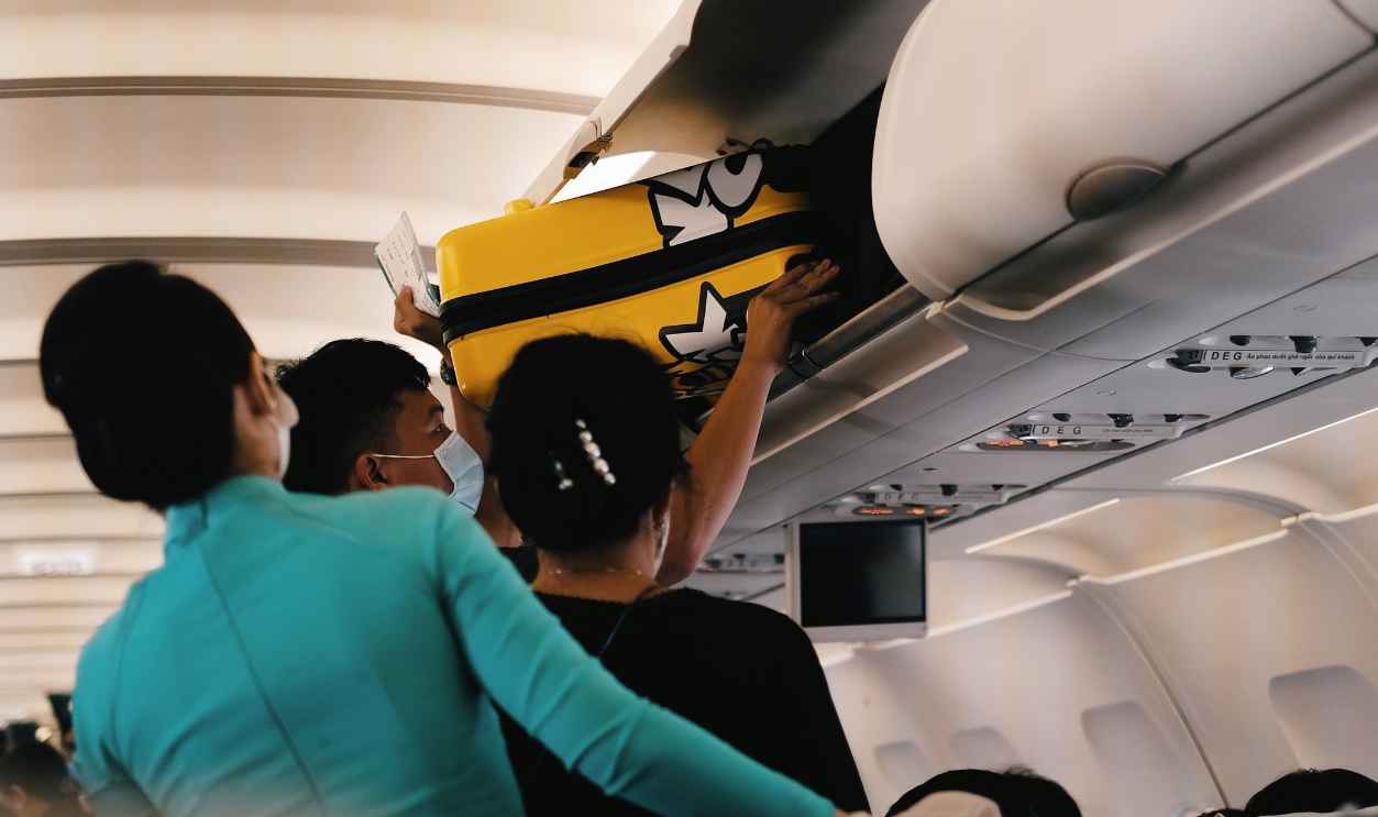 Passengers placing a yellow suitcase in the overhead bin of an airplane cabin.