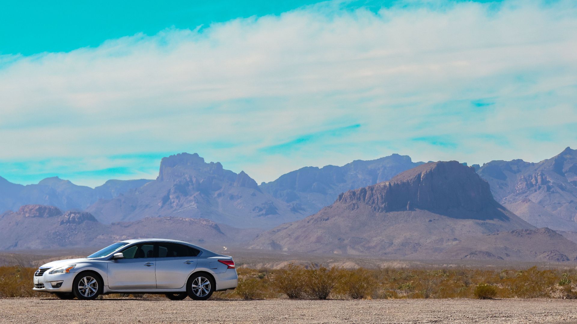 Silver car parked in open desert landscape with striking mountain backdrop under clear blue sky.