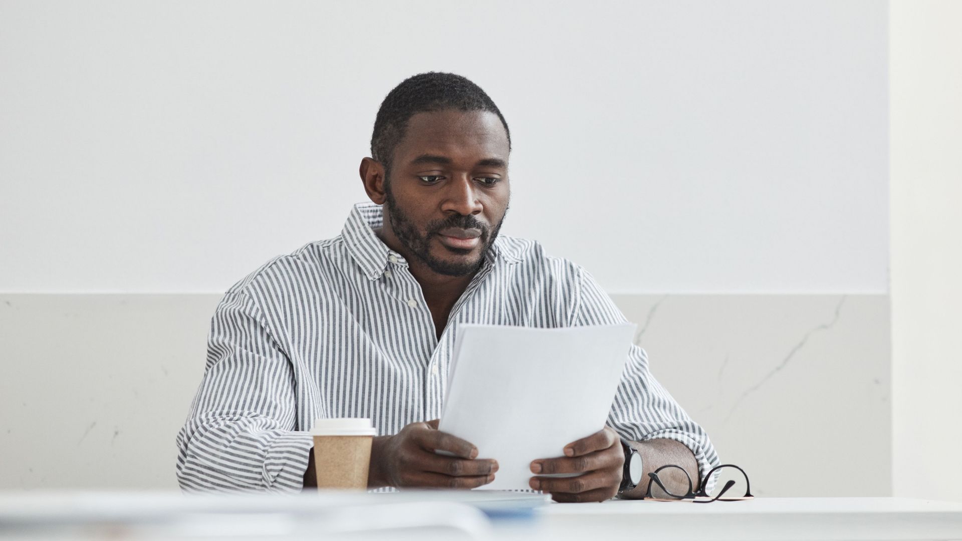 African American man sitting indoors, reading papers with a coffee cup nearby.