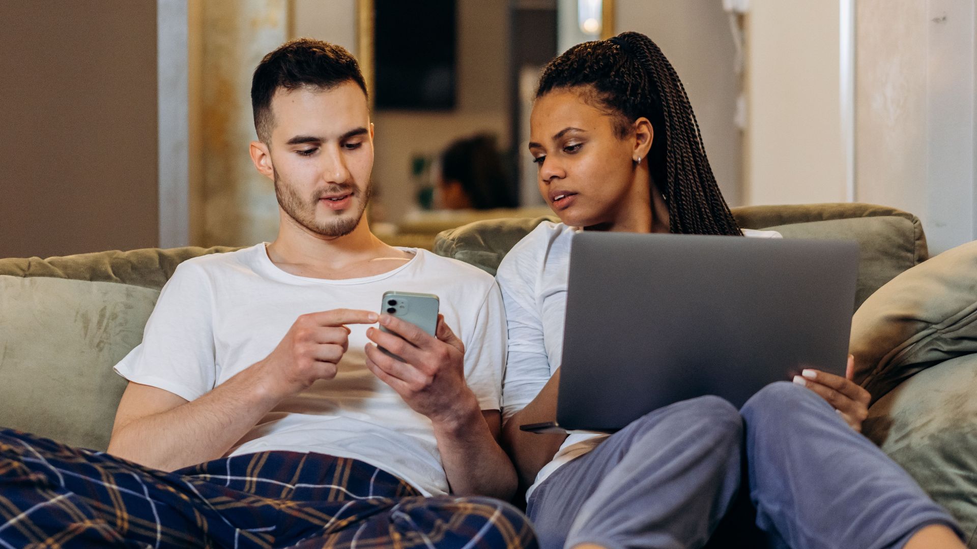 A couple sitting together on a couch, using a laptop and smartphone, enjoying a cozy moment.