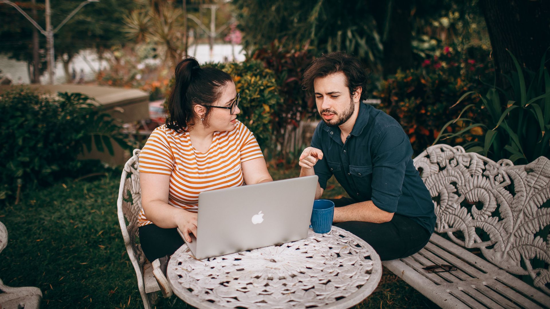 A man and woman working together on a laptop outdoors in a lush garden.
