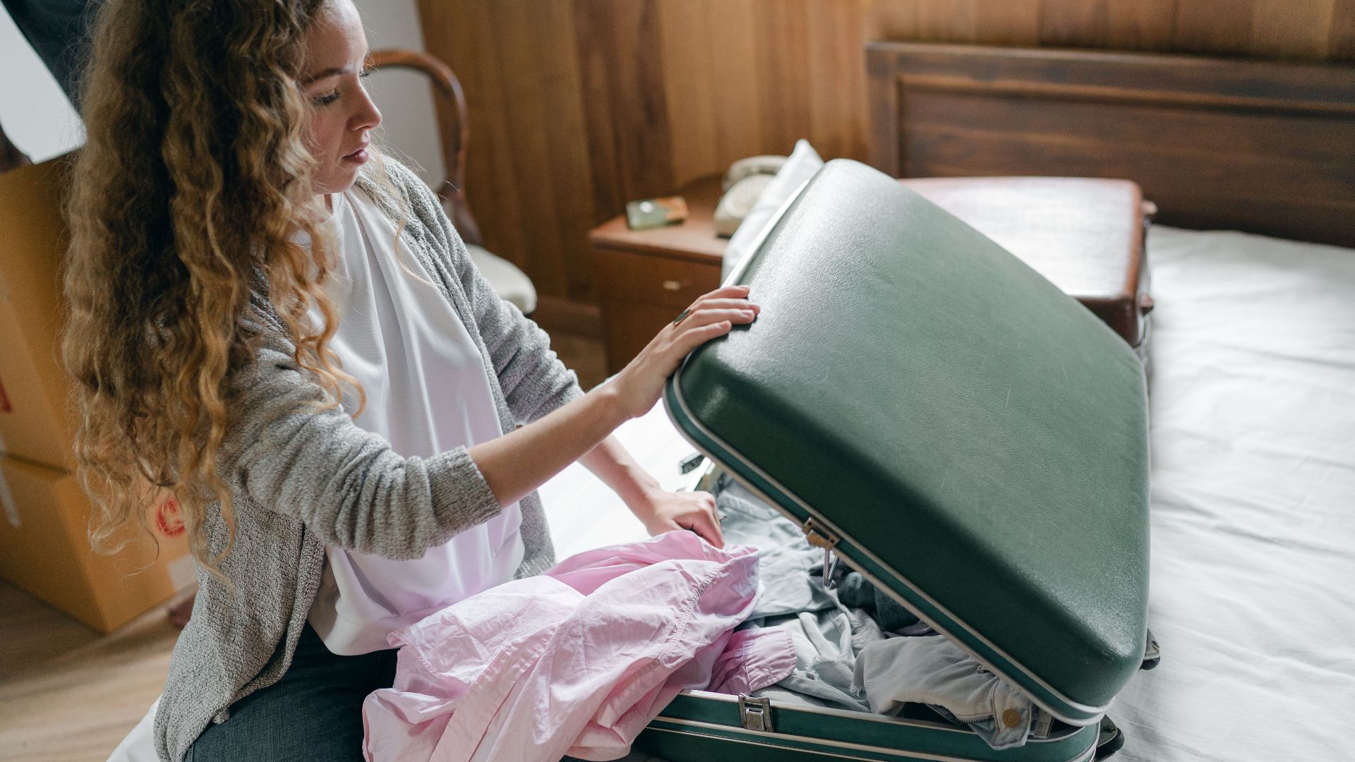 From above concentrated young female in casual wear with long curly hair sitting on bed and packing suitcase before trip