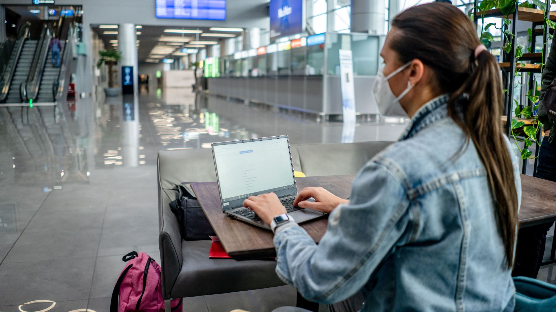 Side view of concentrated female freelancer in protective mask typing on netbook while sitting at table in modern airport terminal