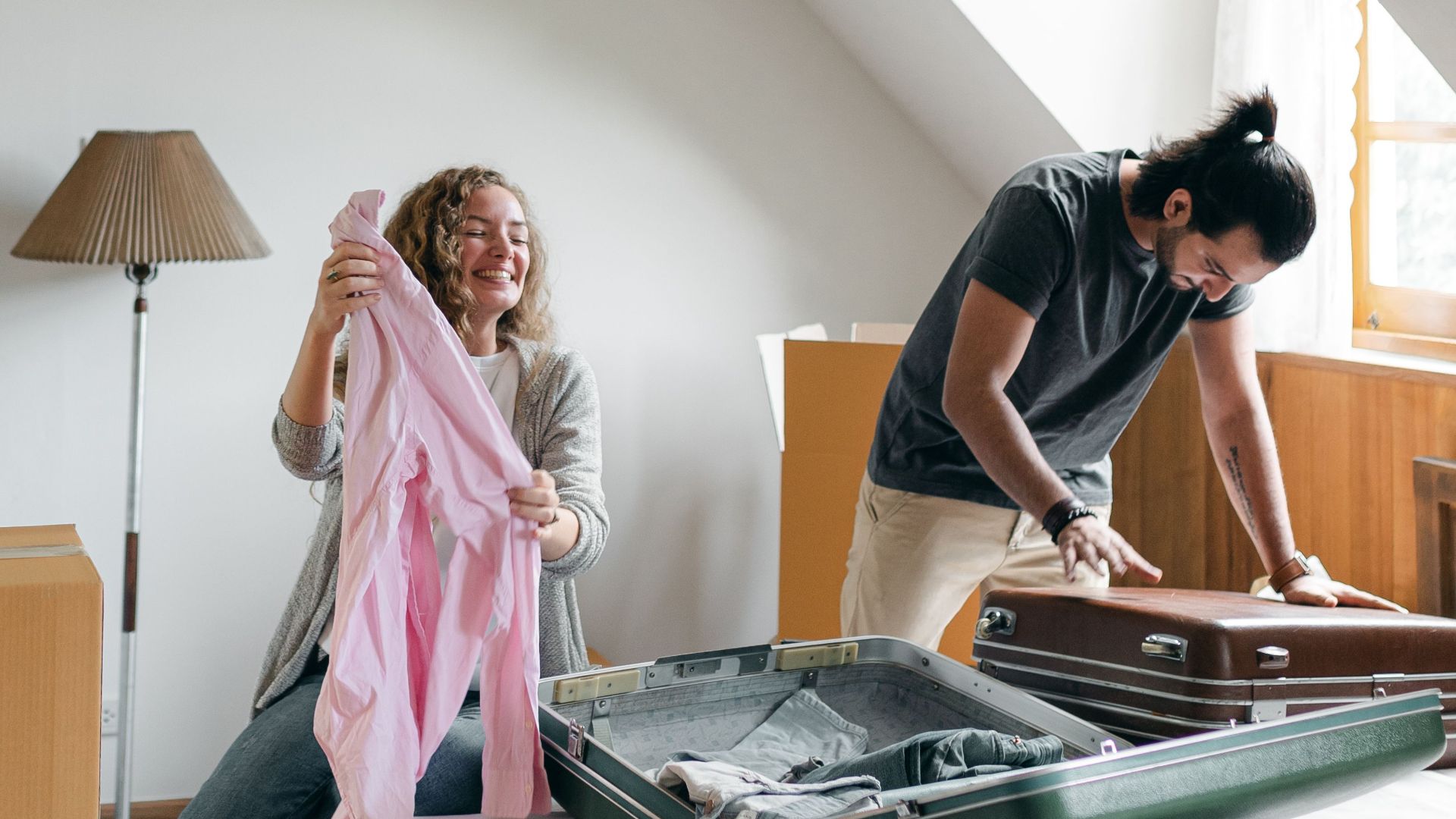 Full body happy young couple packing suitcases and carton boxes while preparing to move into new apartment in empty bedroom on sunny day