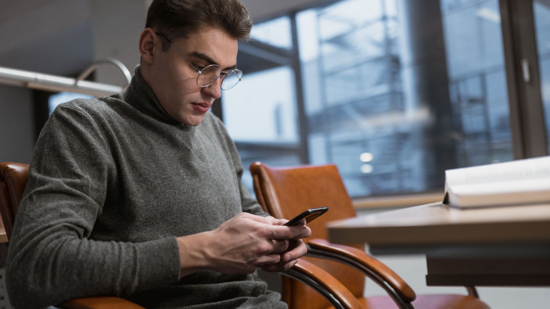 A young man in a grey sweater using a smartphone at an office desk.