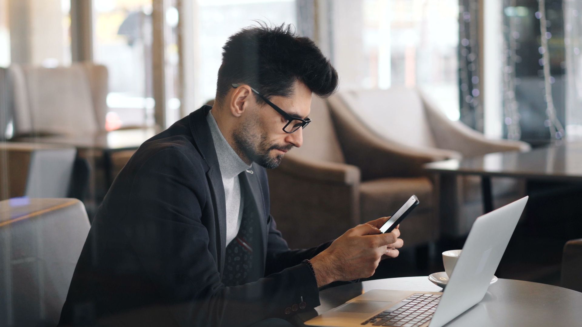 Man in formalwear using smartphone and laptop in a modern café setting.