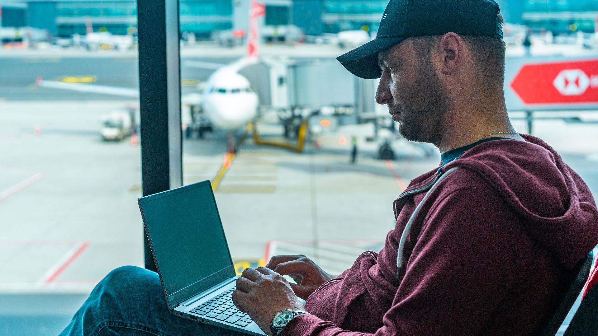 A man working on his laptop in an airport terminal, with a plane visible outside.