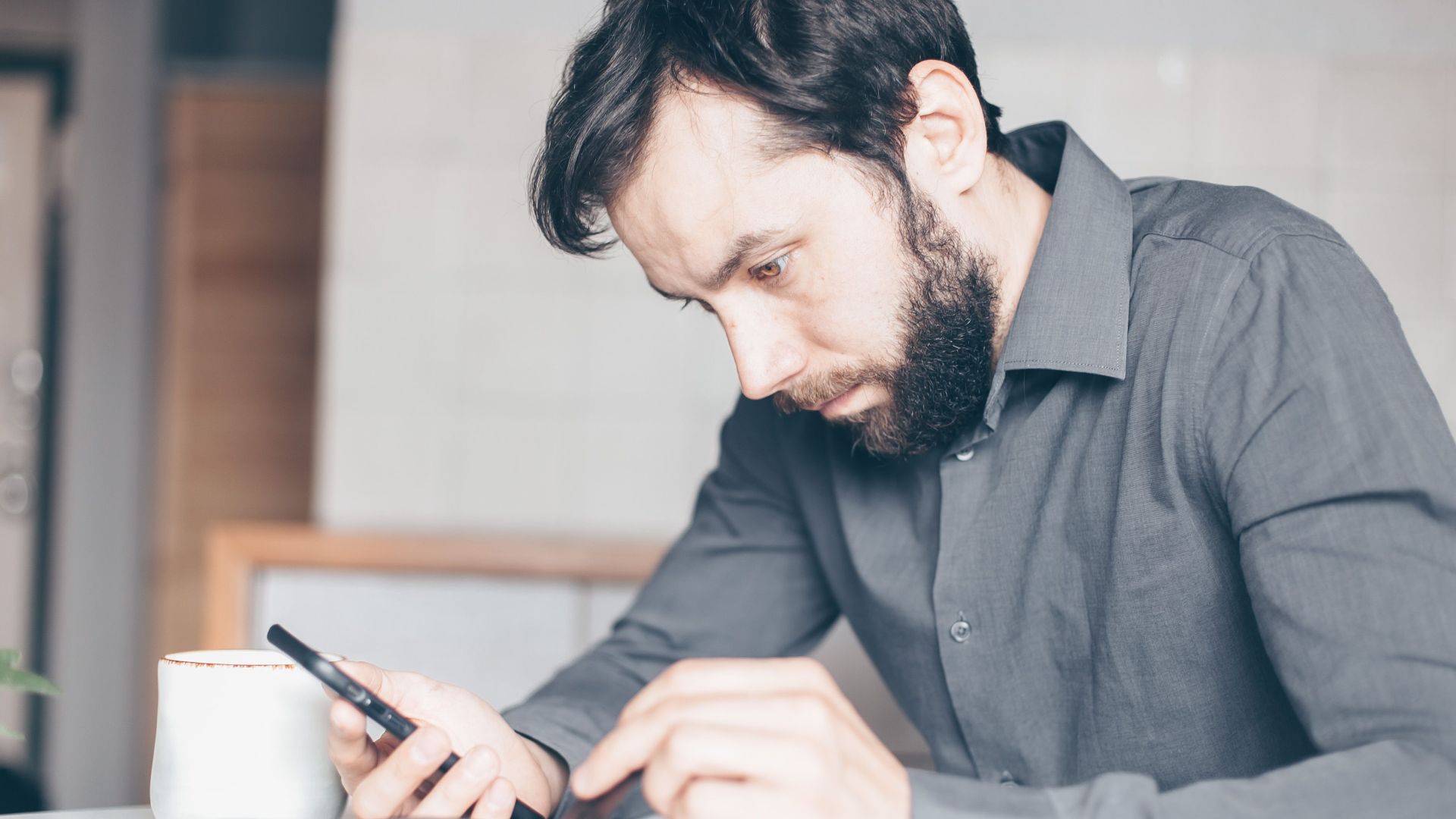 A bearded man intensely stares at his smartphone, deep in thought, indoors.