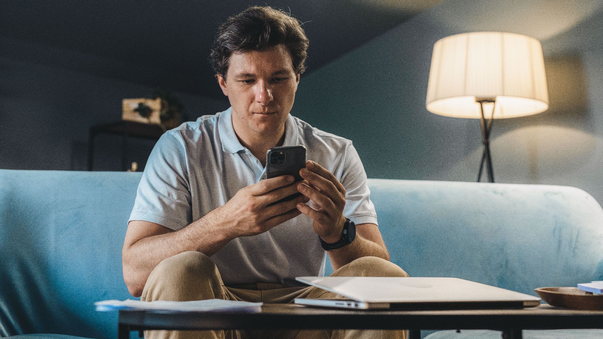 A man sitting on a sofa indoors using a smartphone under warm lighting.