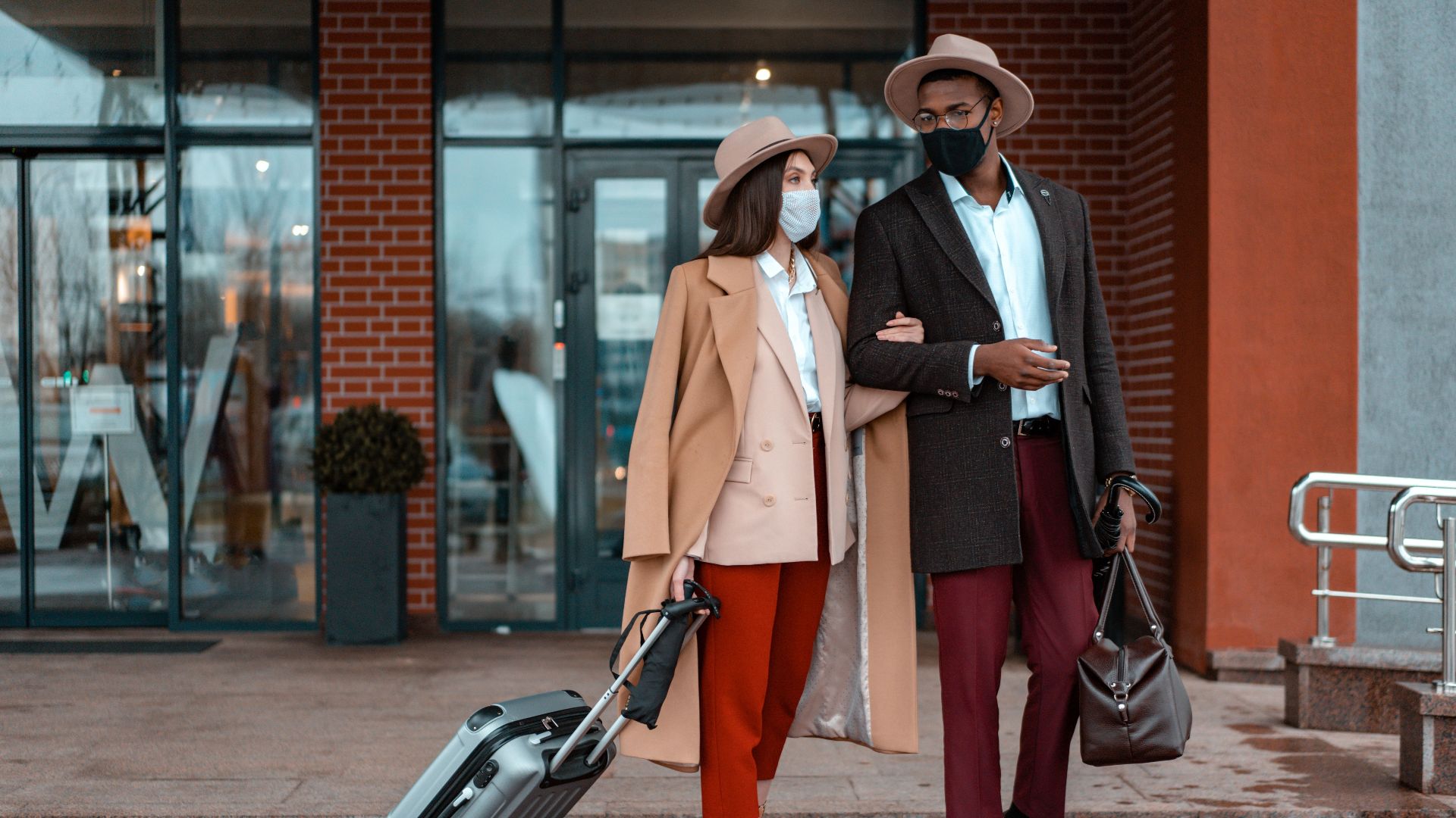Fashionable couple with face masks at hotel entrance, showcasing travel during pandemic.