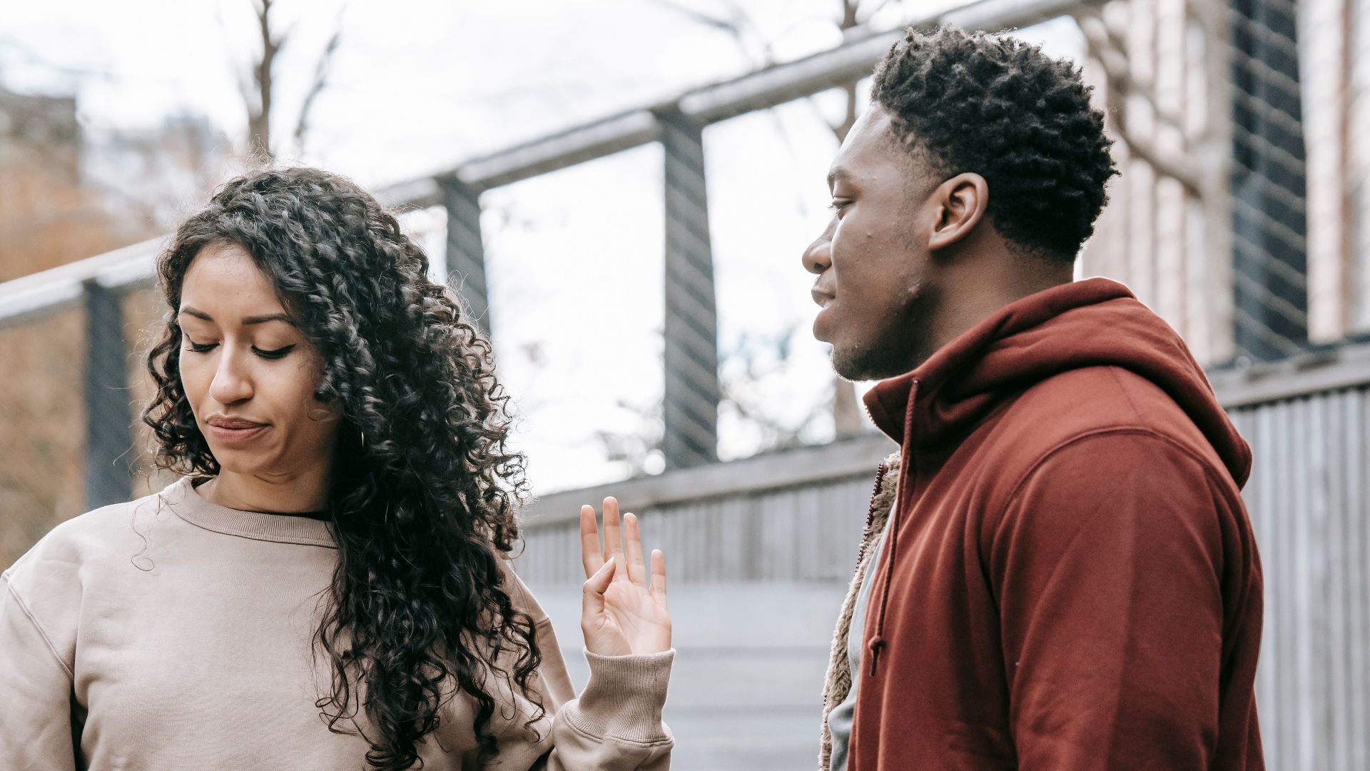Upset ethnic girlfriend with raised hand and concerned African American boyfriend breaking up on street against metal grid on background