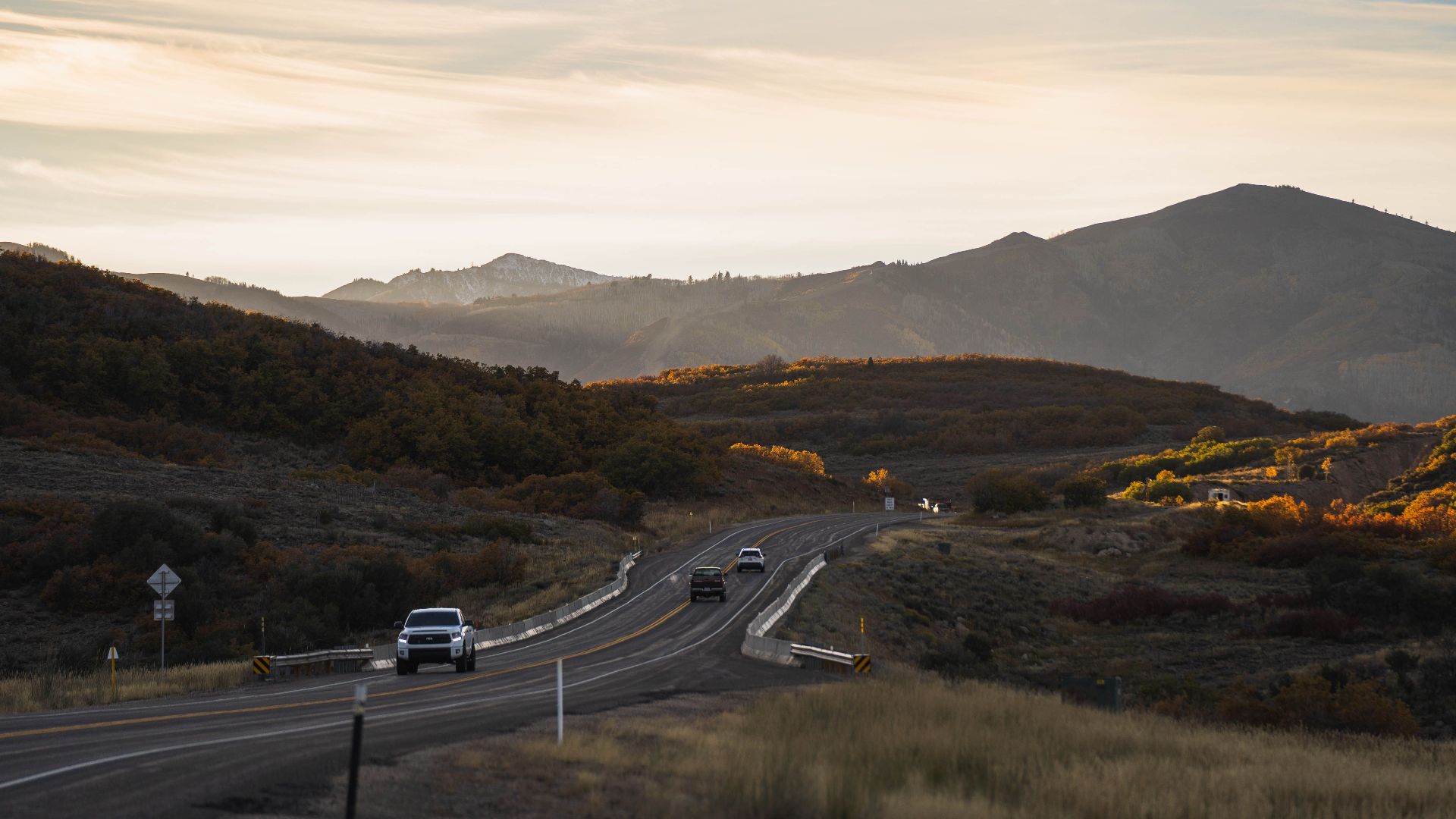 Cars travel on a winding highway through a mountainous landscape during an autumn sunset.