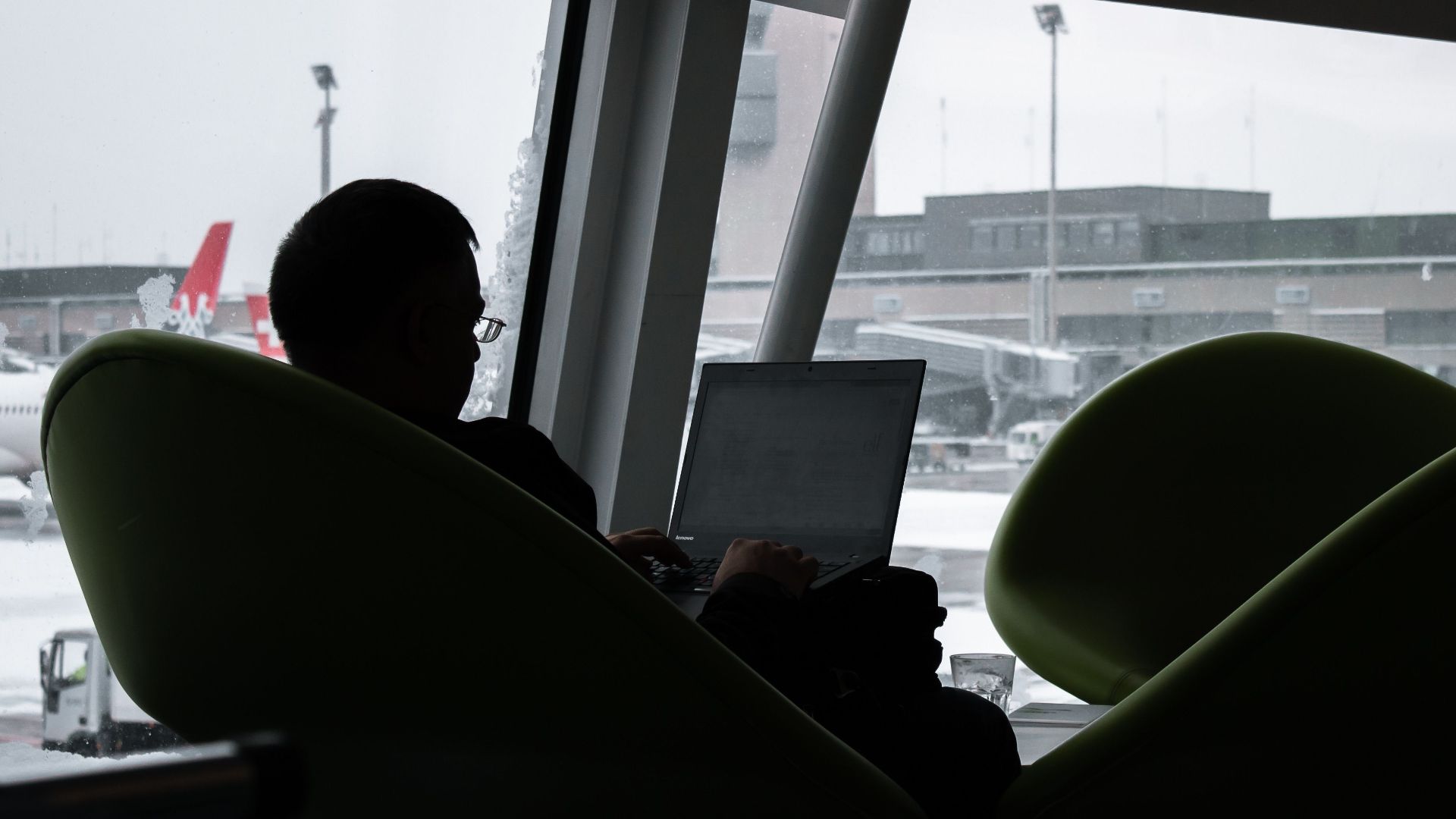 Silhouette of a man working on laptop at airport lounge with planes outside.
