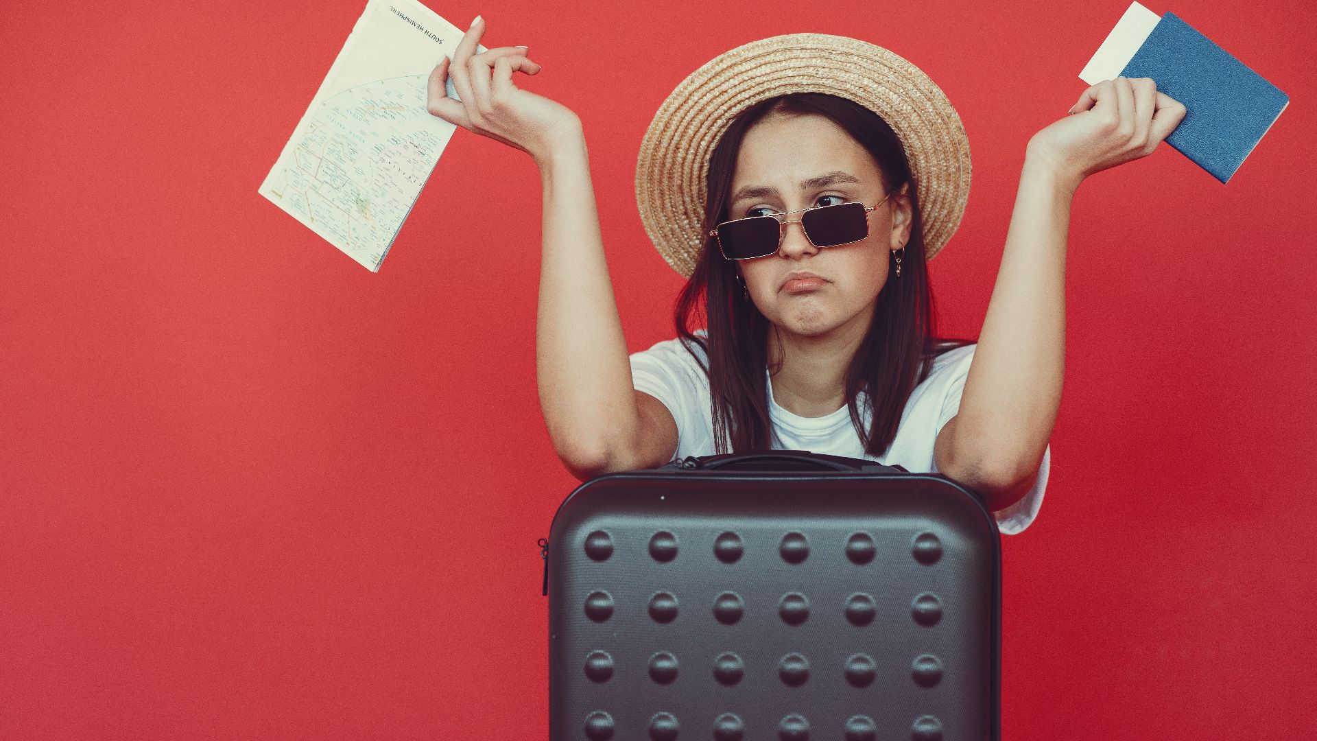 Doubters young stylish woman in wicker hat and trendy sunglasses sitting behind black suitcase and shrugging with passport and tickets on red background
