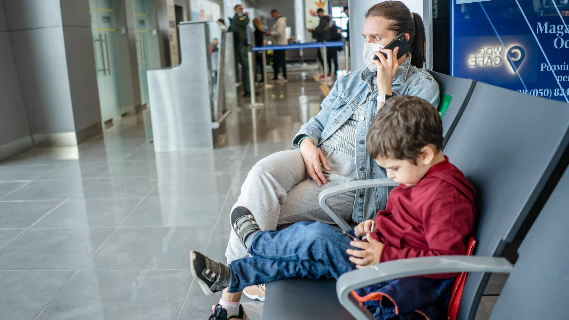 A mother and child sitting at an airport terminal. The mother is on the phone while the child is using a device.
