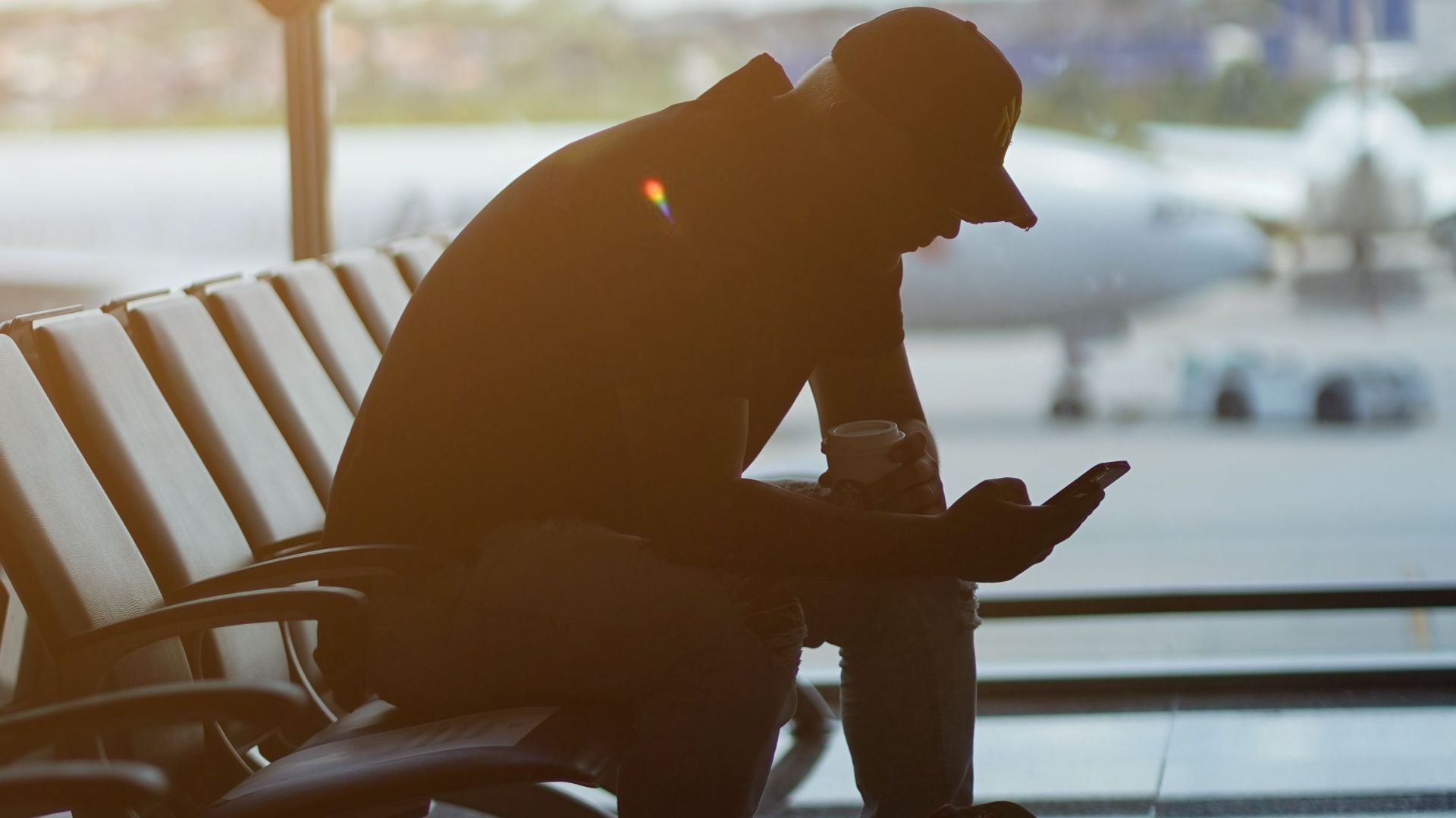 Silhouette of a man using a smartphone in an airport waiting area with airplanes visible outside.
