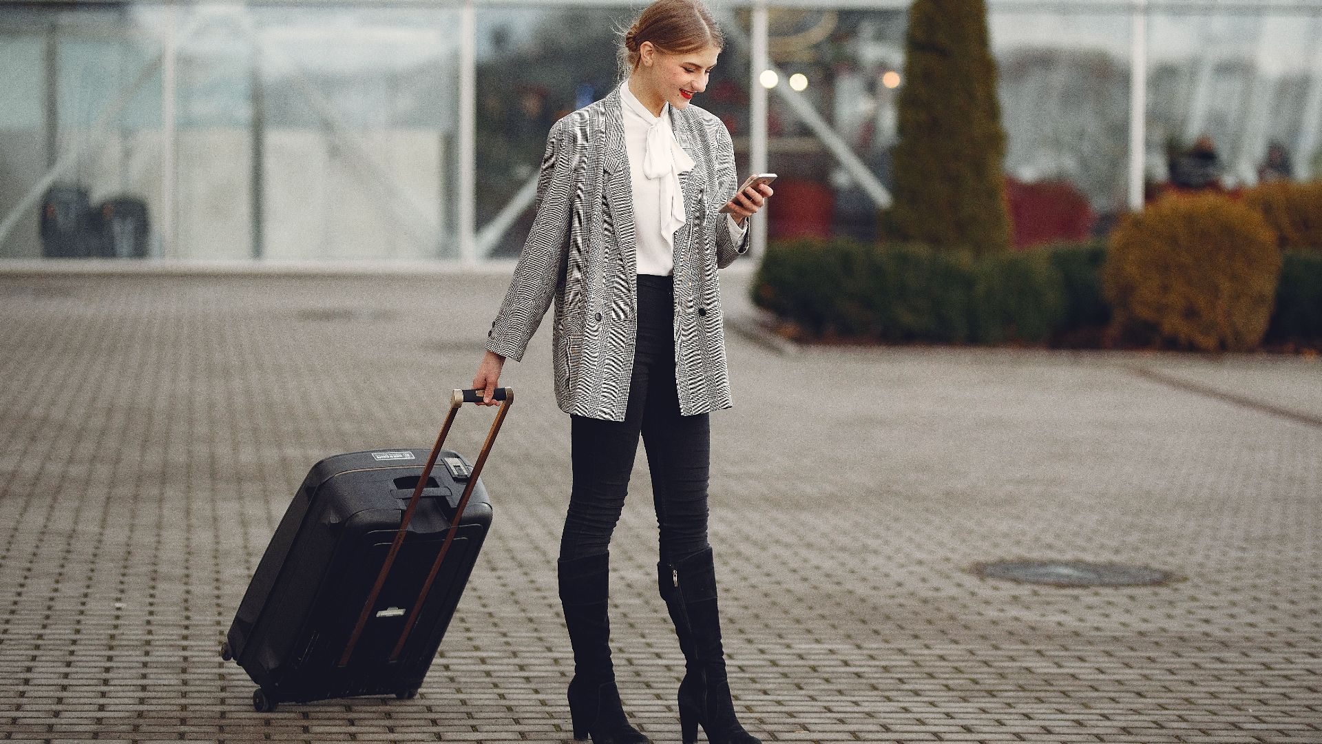 Full length of stylish woman in elegant outfit carrying baggage and surfing smartphone while standing near glass wall of airport