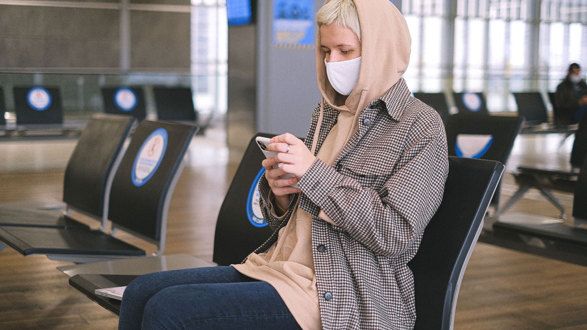 A woman wearing a face mask and a hood sits in an airport waiting area, using her smartphone.