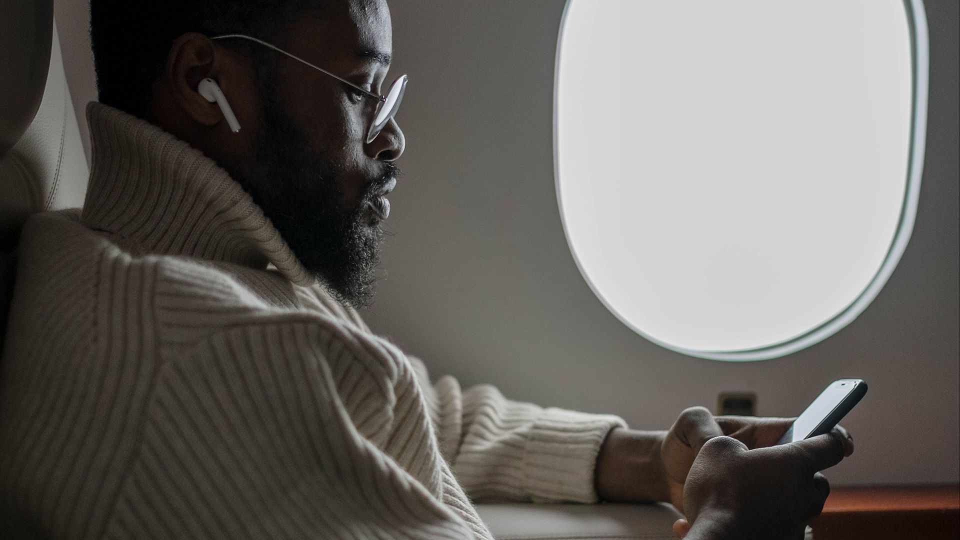 African American man using a smartphone by airplane window, wearing a sweater and glasses.
