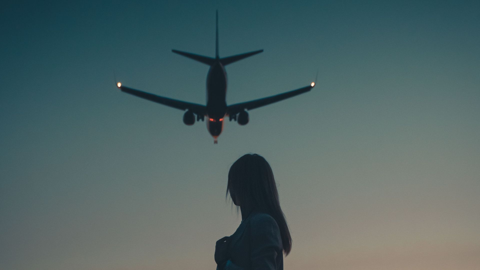 A silhouette of a woman standing as an airplane flies overhead during dusk near an airport.