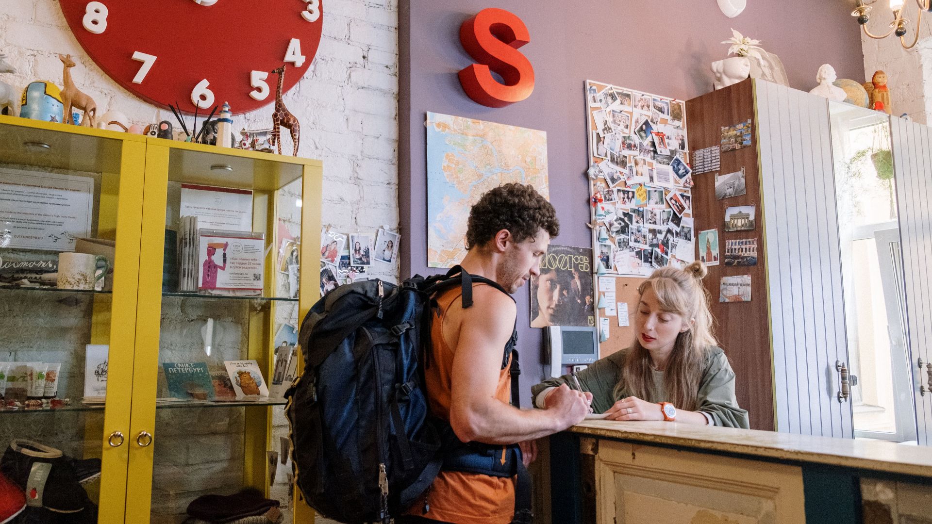 A backpacker at a hostel front desk getting assistance from a receptionist.