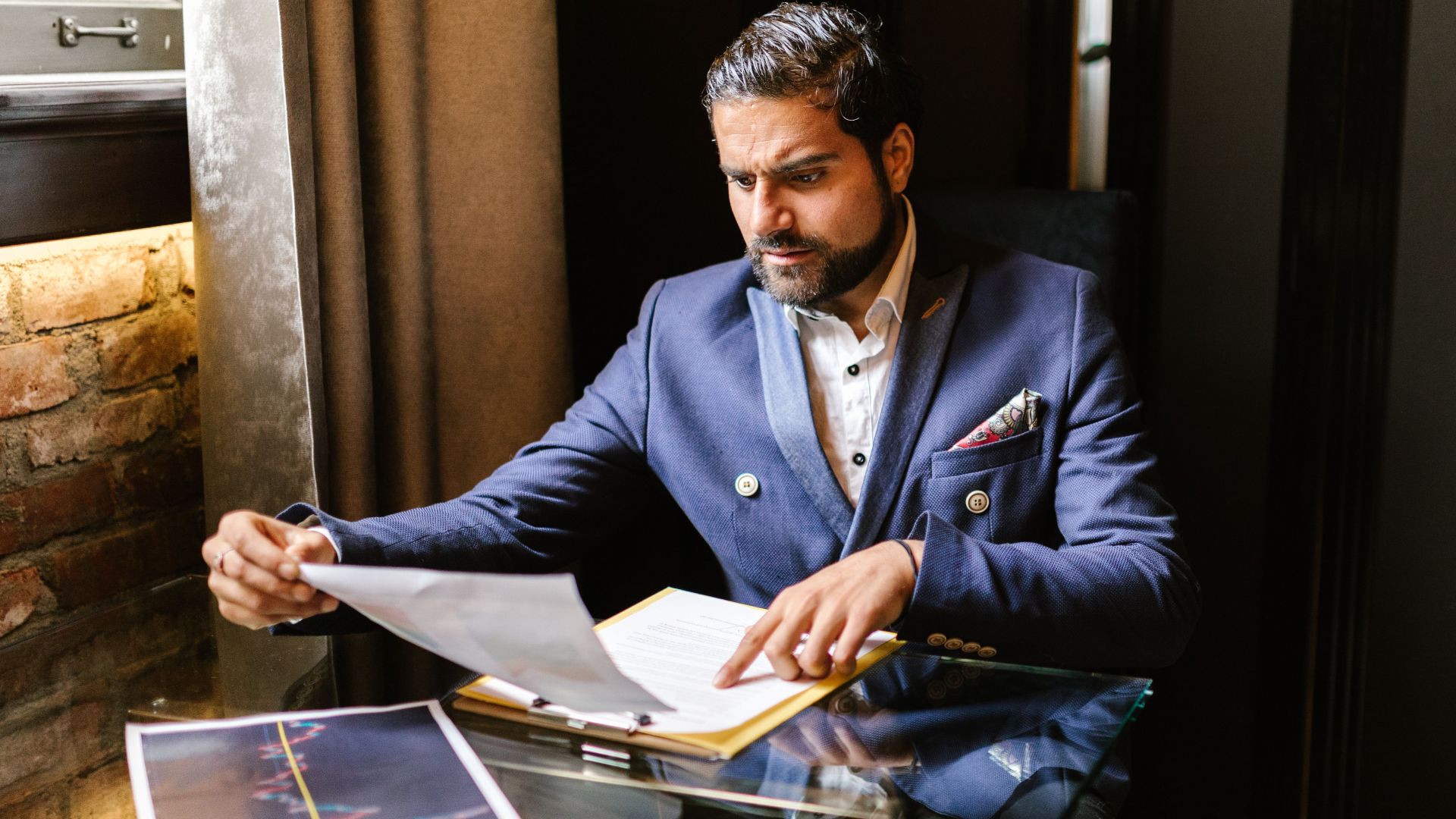 Businessman in blue blazer reviewing financial documents at a glass table.