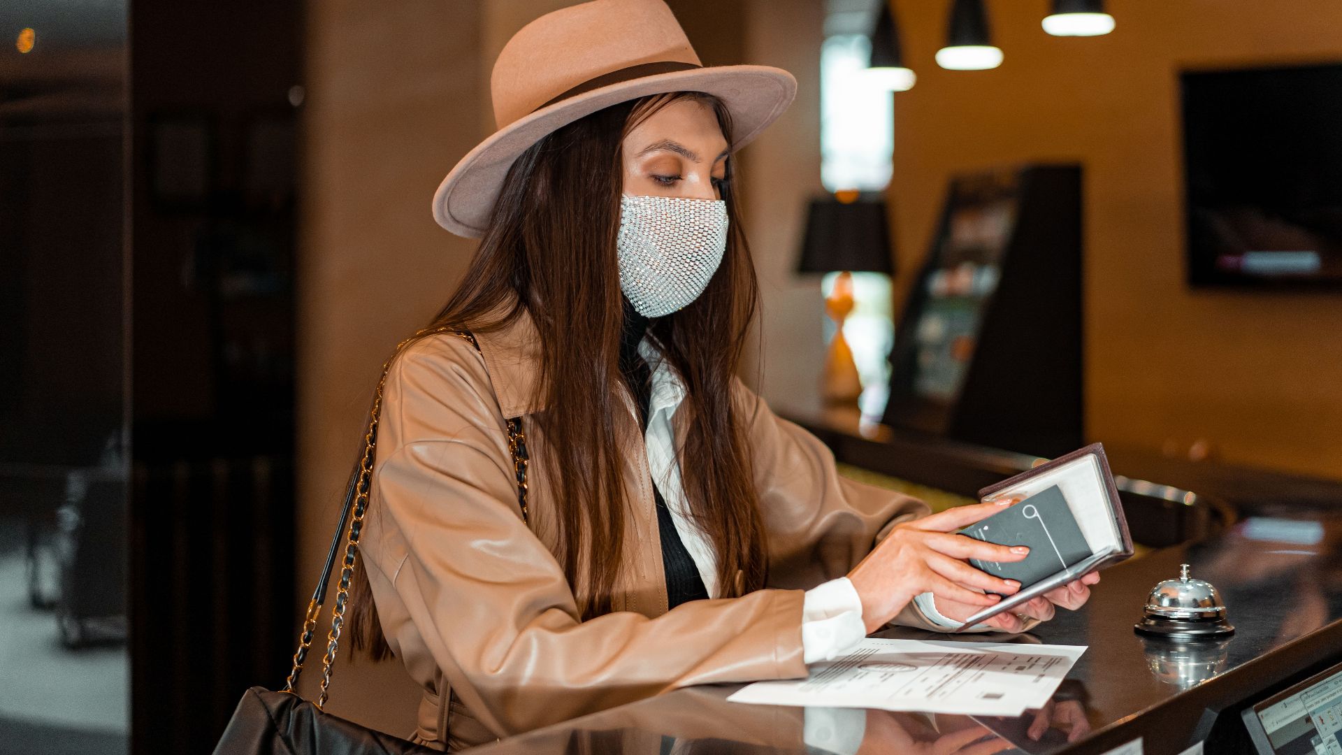 Woman wearing mask and hat checking in at hotel front desk, symbolizing travel and safety.