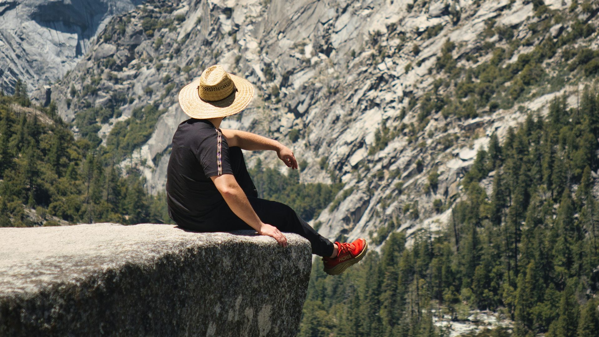 Person in hat sits on cliff overlooking vast mountain valley.