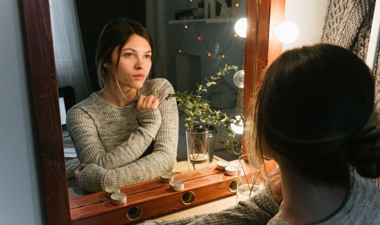 A woman in a cozy sweater contemplates her reflection in a warmly lit room