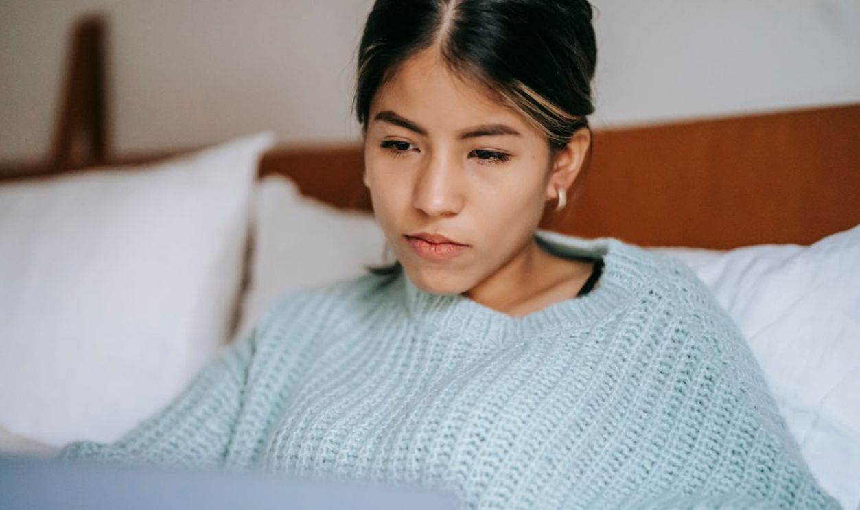 Woman in a sweater focused on her laptop while sitting on a bed, embracing a relaxed indoor atmosphere