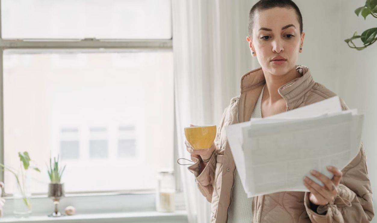 A woman stands by a window holding papers and a coffee cup, in daylight