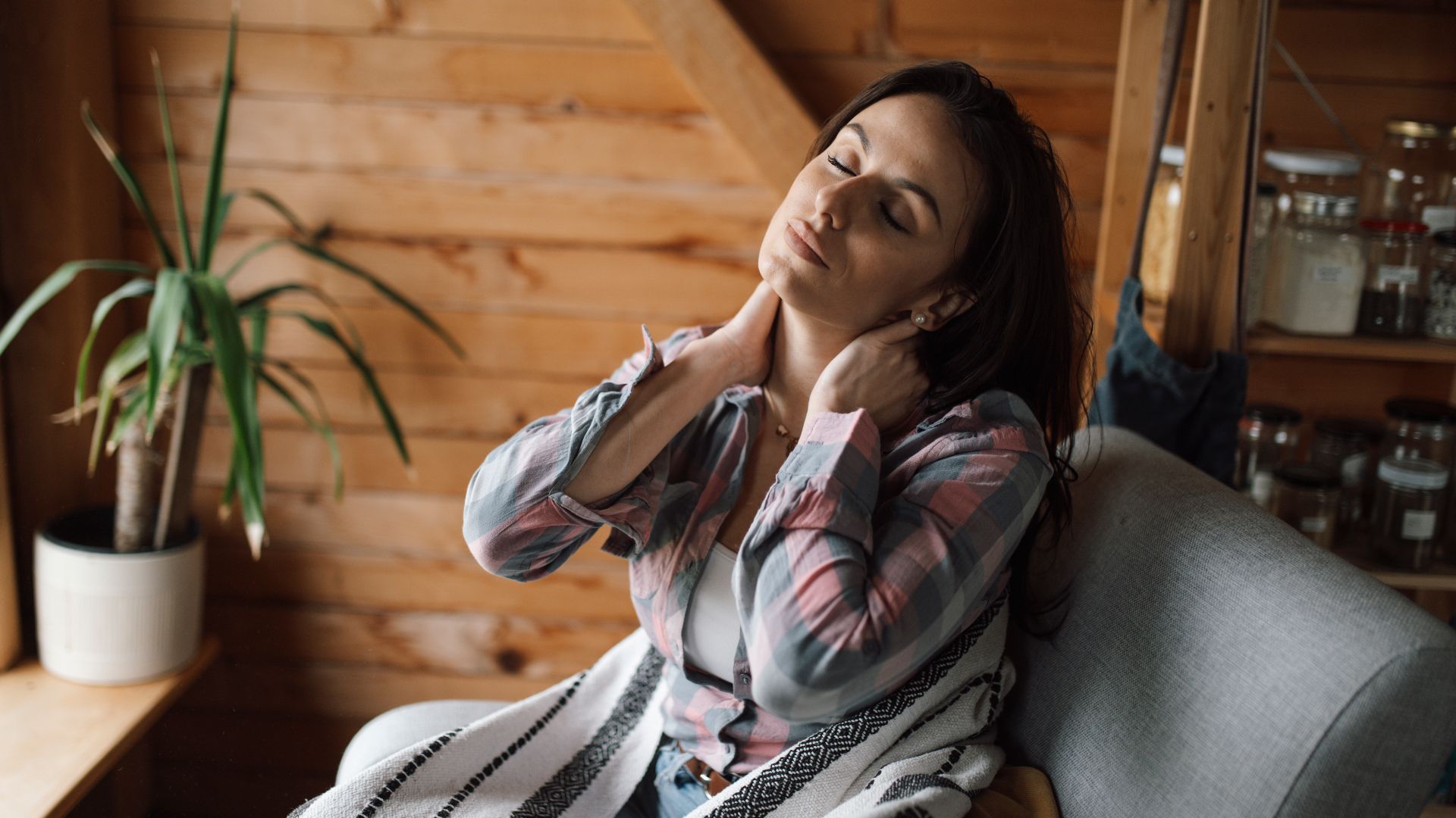 A woman sits on a couch in a wooden room, holding her neck. She seems to be taking a moment to relax and unwind. A plant is placed on the window sill nearby.