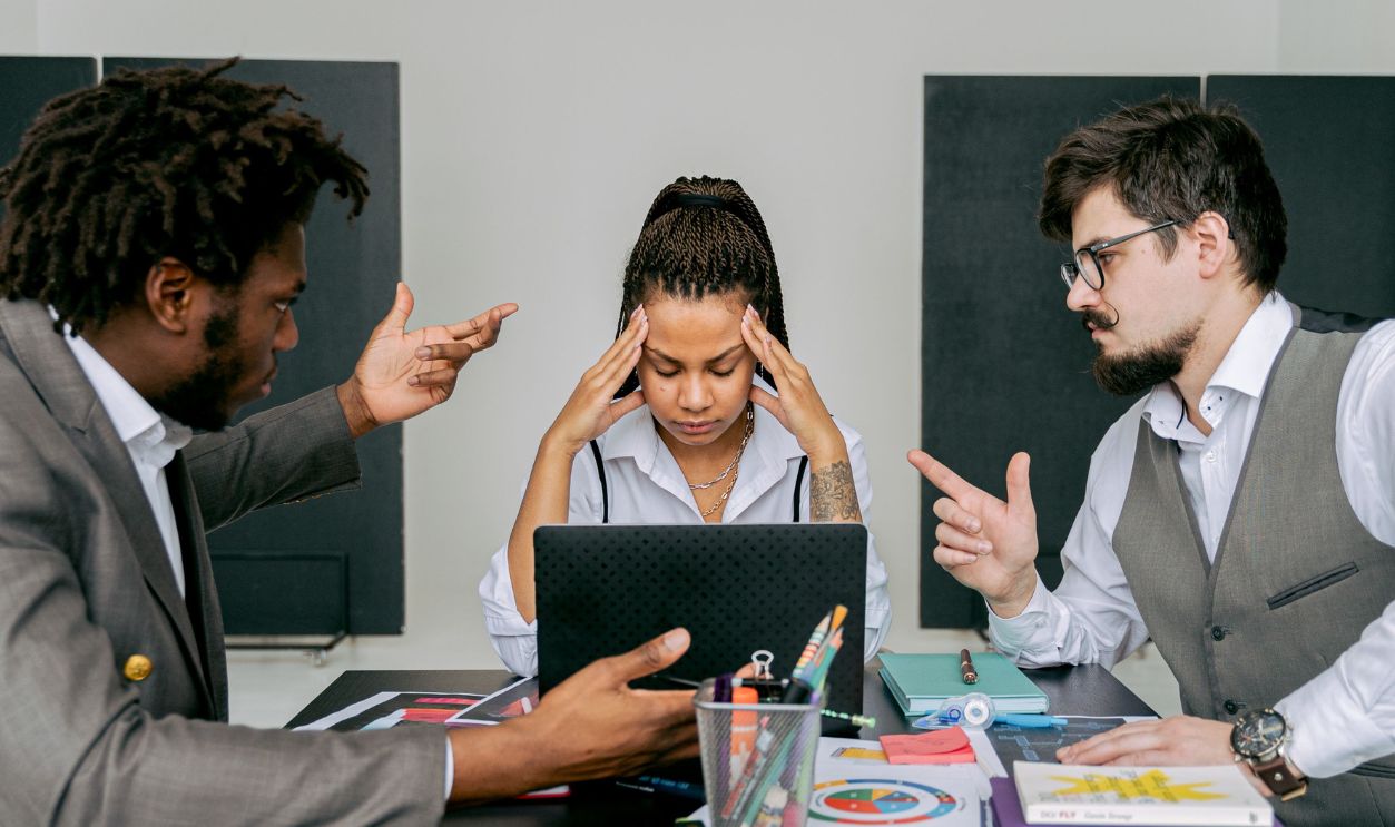 Two men argue while a woman looks frustrated at a laptop in an office