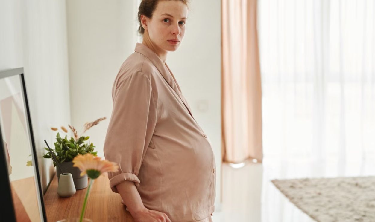 A pregnant woman wearing sleepwear leans on a wooden drawer in a bright, serene room