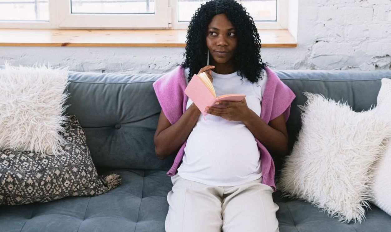 Pregnant woman with a notebook, sitting on a cozy sofa, looking away pensively indoors