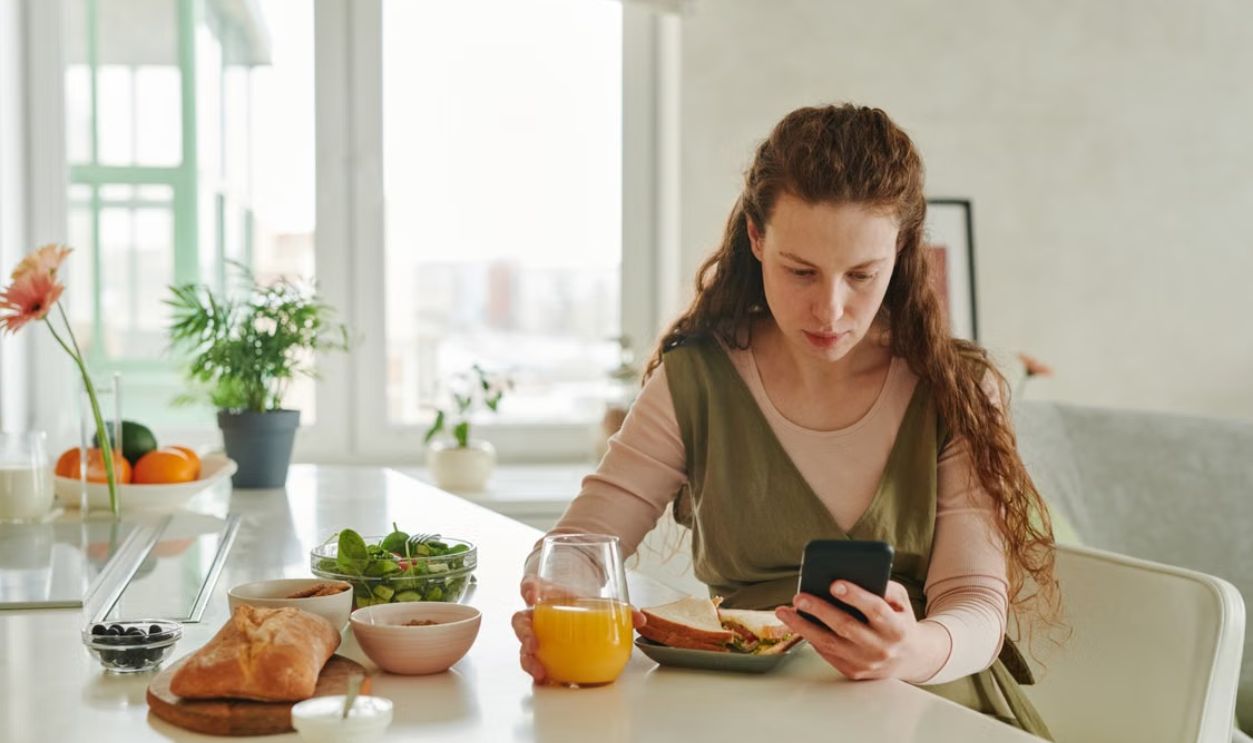 Pregnant woman at breakfast table browsing on smartphone while having a healthy meal indoors.