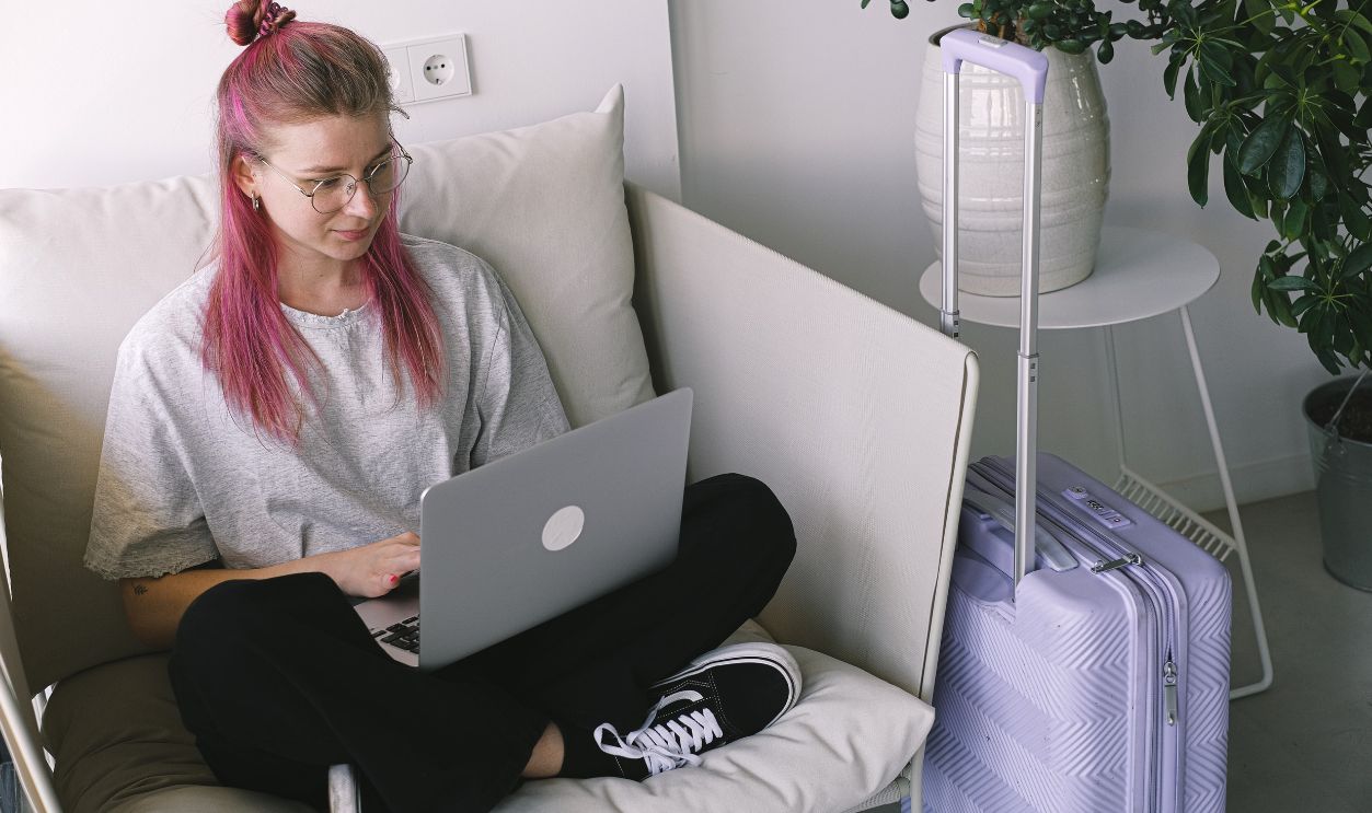 Casual young woman with pink hair works on laptop in comfortable indoor setting, travel suitcase nearby