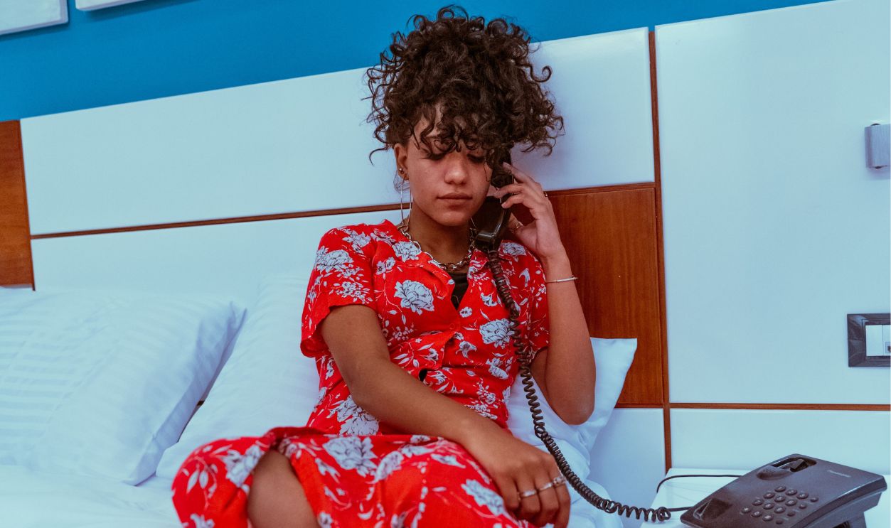 Young woman in vibrant red dress talks on hotel phone while sitting on bed