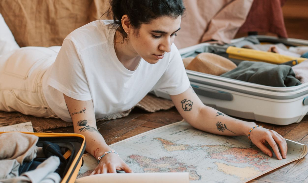 Tattooed woman in white shirt studies map near suitcase on wooden floor indoors. Travel preparation ambiance