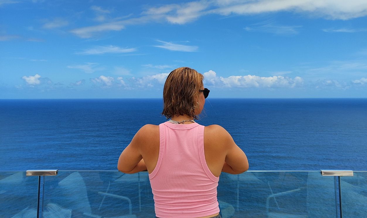 Woman in pink top stands on a glass balcony overlooking the ocean 