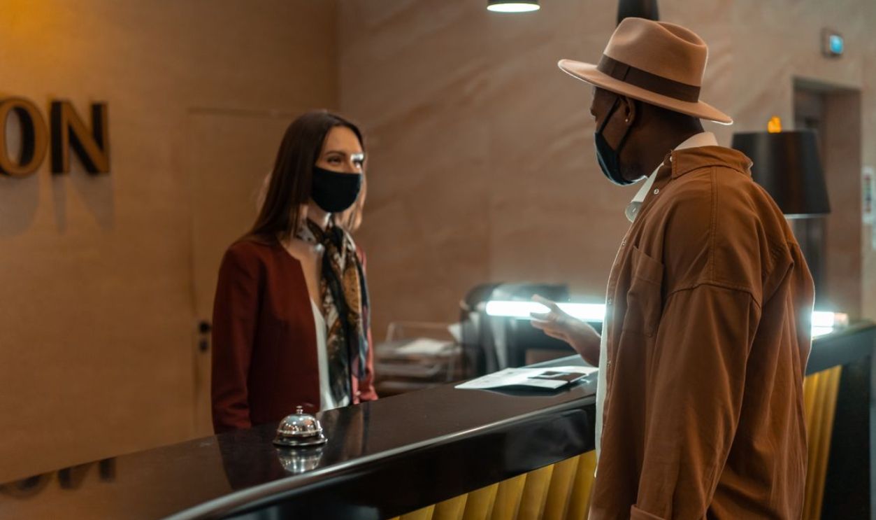 A man at a hotel reception talking to a staff member. Both wear face masks