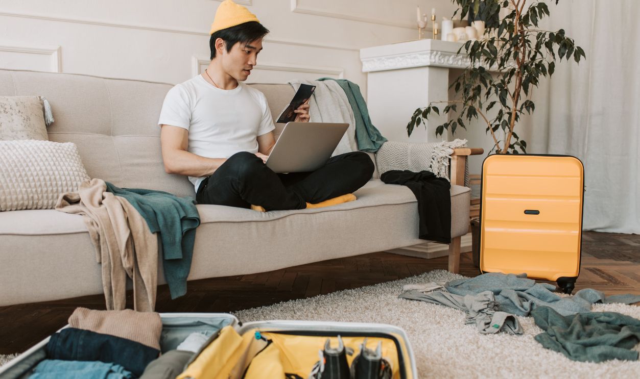 Adult male traveler planning trip at home with laptop and smartphone, surrounded by luggage