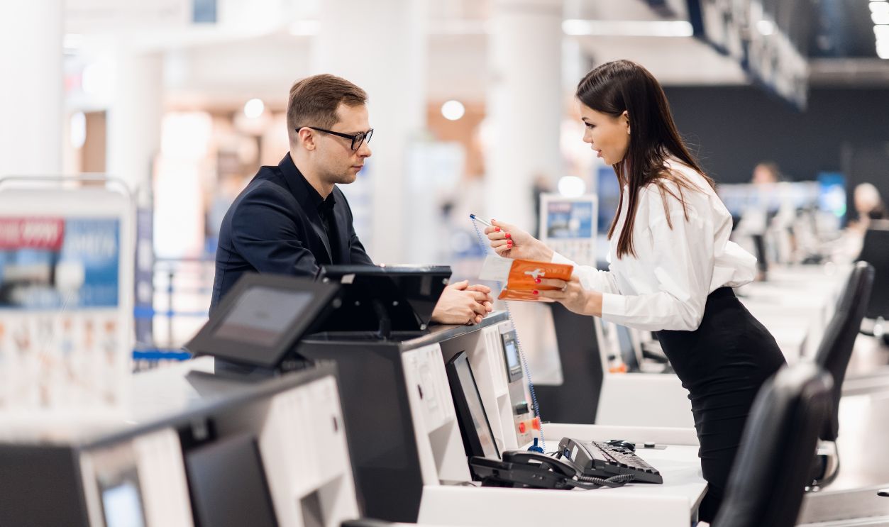 Shutterstock-1609728196, Business trip. Handsome young businessman in suit holding his passport and talking to woman at airline check in counter in the airport